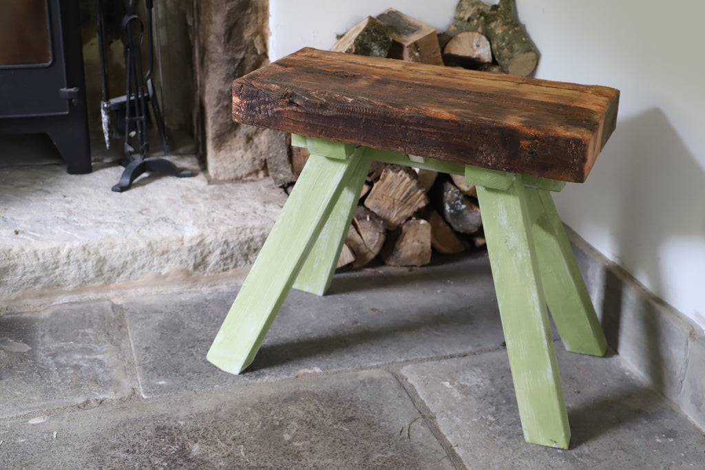 Wooden stool with green legs in front of a fireplace with logs.