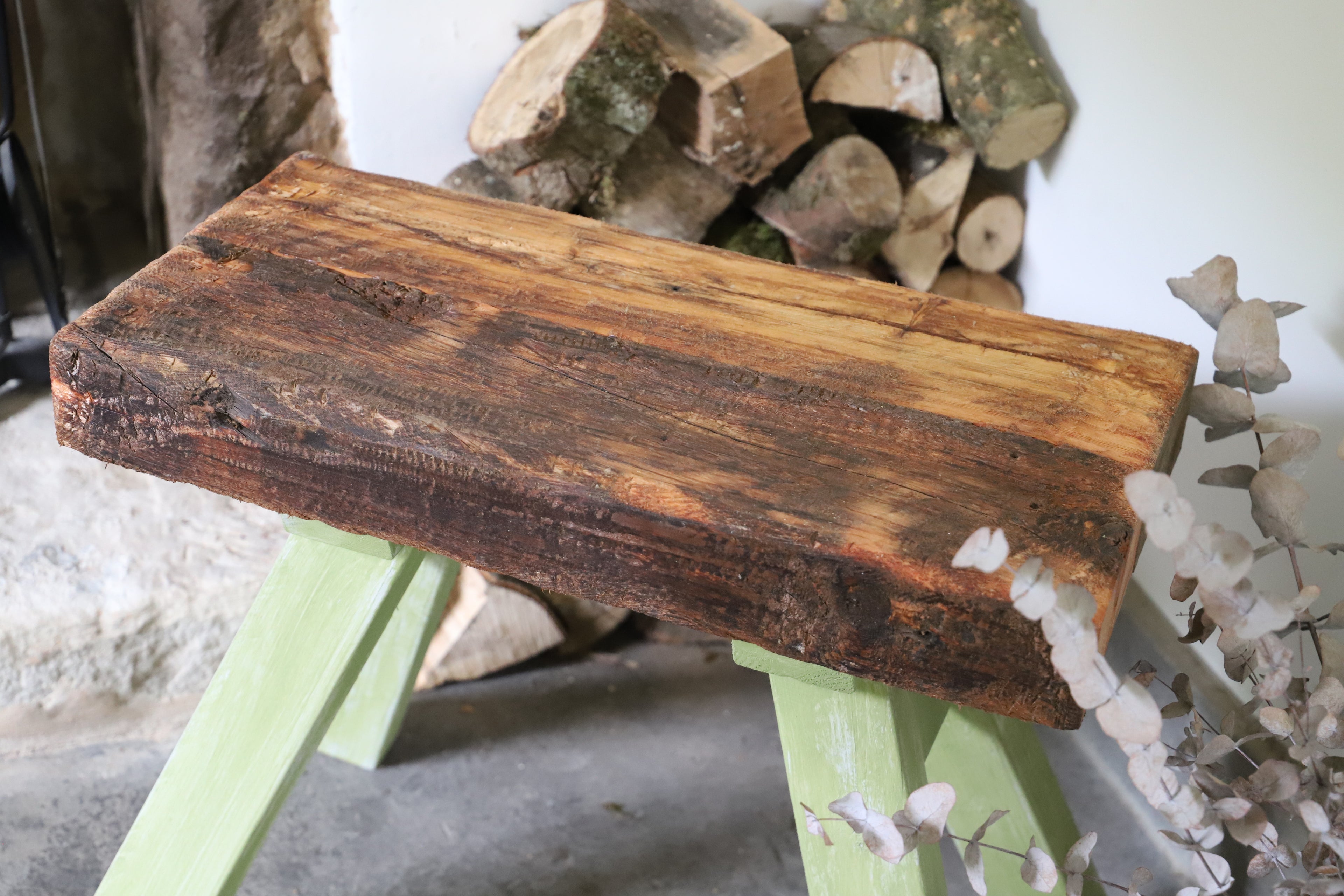 Wooden stool with a rustic finish on a green stand against a background of stacked logs.